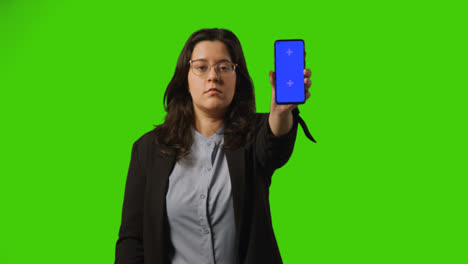 Serious-Young-Businesswoman-Holding-Blue-Screen-Mobile-Phone-Towards-Camera-Standing-Against-Green-Screen-Background